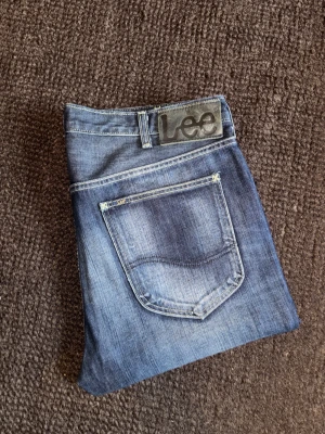 Lee HD Cash slim fit (34) - Mycket snygga blå jeans från Lee modellen Cash, med slim straight fit och coola slitningar. Jeansen  är tillverkade i 100% bomull och har ingen stretch.. Upplagda till benlängd L30 och märkta som W36 men passar bättre för dig på med W34. Mycket fint skick.