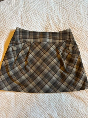 Rutig kortkjol från H&M i grå toner - Supersnygg rutig kortkjol från H&M i olika nyanser av grått och beige. Kjolen har bred linning, dekorativa knappar framtill och dragkedja bak. Perfekt för dig som gillar en klassisk men trendig stil.