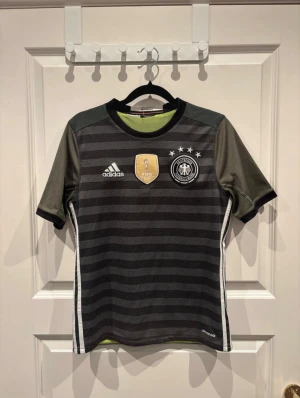 Svart/grön randig Tysklands fotbollströja - Säljer en officiell Tysklands landslagströja från Adidas med svarta och gråa horisontella ränder, korta ärmar och vita sidoremsor. Tröjan har FIFA-märke och landslagsemblem på bröstet samt meshfoder i limegrönt. Materialet är lätt och andas, perfekt för match eller träning. Pris kan diskuteras!!!!