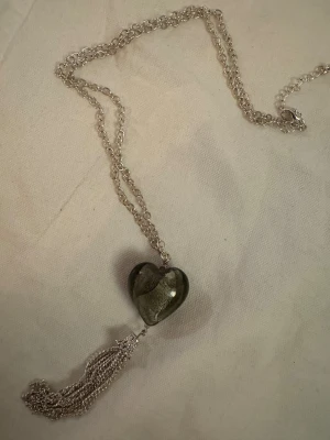 Silverfärgat halsband med hjärthänge - Unikt halsband med silverfärgad kedja och ett stort hjärthänge i glas med metallisk effekt. Under hjärtat hänger en dekorativ tofs av tunna kedjor som ger extra attityd. Perfekt för dig som gillar detaljer och vill sticka ut med ett coolt smycke.