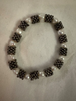 Pärlarmband med silverfärgade detaljer - Unikt armband med oregelbundna vita pärlor och coola silverfärgade kluster mellan varje pärla. Armbandet har en rund form och ger en modern och stilren vibe. Perfekt för dig som gillar att sticka ut med accessoarer som har lite edge.