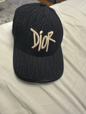 Svart Dior keps med broderad logga - Säljer en svart keps från Dior med broderad vit logga framtill. Kepsen har ett diskret mönster med Dior-monogram över hela ytan och justerbar rem baktill. Materialet är mjukt och känns lyxigt, perfekt för dig som vill sticka ut med en designeraccessoar.
