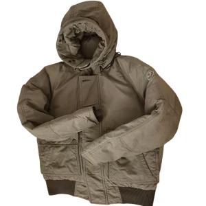 Grön Moncler bomber pufferjacka med huva - Snygg grön pufferjacka från Moncler med stor huva och ribbade muddar. Jackan har två stora fickor framtill med knappar och en klassisk Moncler-logga på ärmen. Perfekt för kalla dagar och har en cool, sportig vibe.