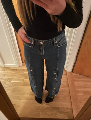 Blå wide jeans med knappar längs benen - Säljer ett par blå jeans med bred passform och knappar längs hela utsidan av benen. Jeansen har klassisk femficksdesign och är tillverkade i robust denim. Perfekt för dig som gillar en avslappnad och trendig stil.
