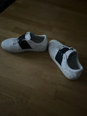 Valentino Garavani vita sneakers - Snygga vita sneakers från Valentino Garavani med bred marinblå rem över mitten och klassisk snörning. Skorna har rund tå, platt sula och är tillverkade i skinn. Perfekt för dig som gillar stilrena designer med en lyxig touch. Vid frågor kom dm 