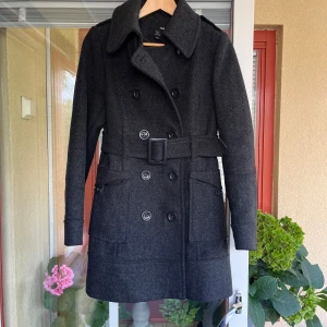 Mörkgrå kappa i ull från H&M - Säljer en mörkgrå kappa från H&M i storlek 34. Den har en klassisk knäppning och ett skärp i midjan. Snyggt foder. Material 80% ull och polyamid 20%. Fodret är polyester. 