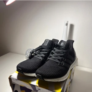Adidas Ultraboost svarta sneakers - Svarta Adidas Ultraboost sneakers med stickad ovandel och klassiska tre ränder på sidan. Skorna har en vit, responsiv Boost-mellansula och svart yttersula med Continental-gummi för extra grepp. Perfekta för dig som gillar stilrena och bekväma sneakers.
