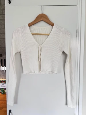 Vit croppad ribbad topp med knytband, storlek M - Säljer en vit croppad topp med lång ärm och snygg v-ringning. Ribbad och har knytband framtill. Stilren och enkel. Materialet är mjukt och är mycket stretchigt. Finns två små fläckar på baksidan av vänster ärm som inte får bort i tvätt. Knappt använd. - Köper du innan kl 16:00 kan jag posta samma dag!