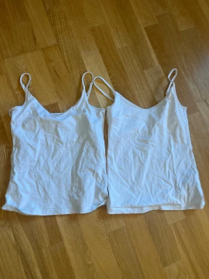 Vita linnetoppar från 157, XS & S - Två vita linnetoppar från 157 med smala axelband och v-ringad hals. Materialet är mjuk bomull med lite stretch (95% bomull, 5% elastan) som sitter skönt mot kroppen. Perfekt att styla med jeans eller shorts för en clean look.