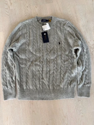 Grå kabelstickad tröja Polo Ralph Lauren - Kabelstickad grå tröja från Polo Ralph Lauren med klassisk rund hals och broderad logga på bröstet. Tillverkad i en mjuk blandning av ull, akryl och nylon. Ribbstickade muddar vid ärmslut och nederkant ger en snygg passform.