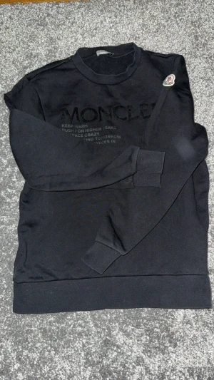 Svart sweatshirt från Moncler - Svart sweatshirt från Moncler med broderad logga på bröstet och patch på ärmen. Tröjan har rund hals, ribbade muddar och är tillverkad i mjuk bomull. Perfekt för dig som gillar stilrena och exklusiva märken.