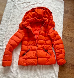 Orange pufferjacka från GANT - Säljer en fet orange pufferjacka från GANT med huva och två dragkedjeförsedda fickor. Jackan har quiltad design och snygga detaljer som logopatch på ärmen. Perfekt för kalla dagar när du vill sticka ut med färg.