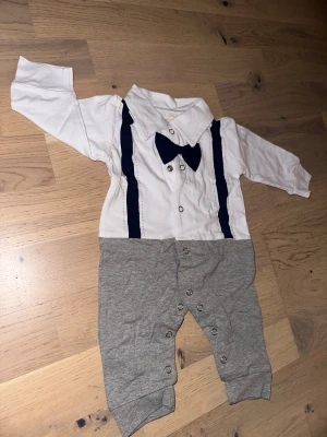 Babykostym med fluga och hängslen Vit/grå body, storlek 70 - Supersöt babykostym med vit skjortdel, mörkblå fluga och hängslen samt grå byxdel. Tryckknappar framtill och längs benen gör den enkel att ta på och av. Perfekt för små stiliga tillfällen!