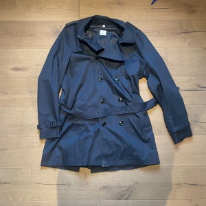 Svart trenchcoat från Burberry, stl 56 - Klassisk svart trenchcoat från Burberry med snyggt bälte i midjan och dubbla knapprader. Jackan har långa ärmar, stilren krage och exklusivt Burberry-foder med logga. Perfekt för dig som vill ha en tidlös och lyxig look.
