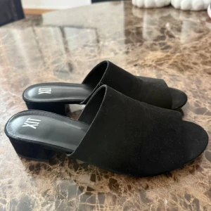 Svarta mockasandaler med klack från XIT - Stilrena svarta sandaler från XIT i mjuk mocka med öppen tå och bred rem över foten. Skorna har en låg klack och platt sula, perfekta för en trendig look. Enkla att matcha till olika outfits och riktigt bekväma att bära.