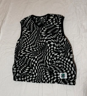 Svartvit fleeceväst med optiskt mönster - Cool svartvit fleeceväst från H&M med hypnotiskt rutigt mönster och v-ringning. Västen har en liten patch nertill med logga och är perfekt för dig som vill sticka ut. Mjuk och skön med en loose fit.