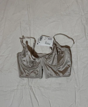 Champagnefärgad satin korsett från H&M - Snygg champagnefärgad korsett från H&M i glansigt satinmaterial. Modellen har smala axelband, markerade kupor och en stilren design med hakknäppning framtill. Perfekt för att lyfta din outfit med en trendig touch.