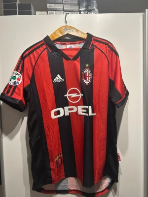 AC Milan Maldini #3 Adidas fotbollströja - Säljer en klassisk AC Milan fotbollströja från Adidas med Maldini och nummer 3 på ryggen. Tröjan har röd-svarta vertikala ränder, korta ärmar, broderade klubb- och Serie A-märken samt Opel-logga på bröstet. Materialet är lätt och ventilerande polyester. Aldrig använt.