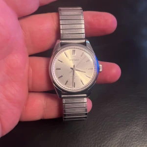 Vintage Klassisk silverfärgad Seiko klocka - Säljer en stilren Seiko klocka med rund boett och silverfärgad urtavla. Armbandet är flexibelt och i metall med horisontella ränder. Klockan har minimalistiska index och en diskret krona på sidan. Perfekt för dig som gillar tidlös design och metalliska detaljer.