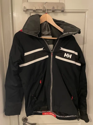 Svart och röd vindjacka Helly Hansen - Säljer en svart vindjacka från Helly Hansen med röd huva och vita detaljer. Jackan har dragkedja framtill, två fickor med röda drag, och reflekterande ränder på bröstet och ärmsluten. Perfekt för blåsiga dagar och har en sportig look med HH-logga på bröstet och ärmen.