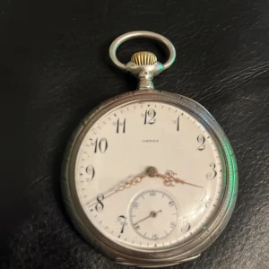 Silver Omega fickur Grand Prix Paris 1900 - Säljer ett klassiskt Omega fickur med rund boett i silverfärgat metall och vit urtavla. Snygga svarta siffror och dekorativa visare i guldton. Insidan har graverad text 'Grand Prix Paris 1900' och detaljerad mekanik i guld och silver. Perfekt för dig som gillar vintage och coola accessoarer.