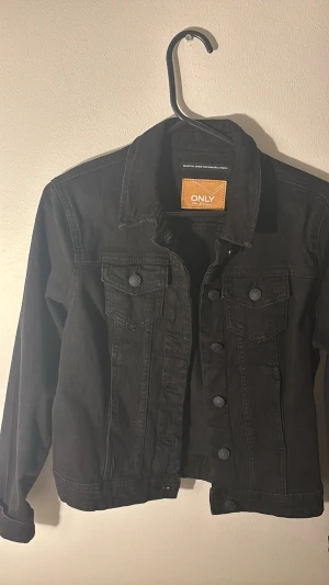 Svart jeansjacka från ONLY - Snygg svart jeansjacka från ONLY med klassisk krage, två bröstfickor med knappar och raka långa ärmar. Jackan har en stilren look med svarta knappar framtill och är tillverkad i slitstarkt denim-material. Perfekt för dig som gillar enkel och cool stil.