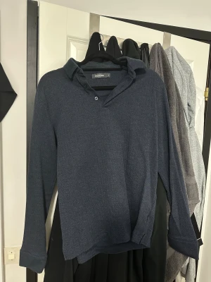 Mörkblå pikétröja från Dressmann - Snygg mörkblå pikétröja från Dressmann med diskret fiskbensmönster. Tröjan har krage, knappslå och långa ärmar. Passar dig som vill ha en chill men ändå dressad look.