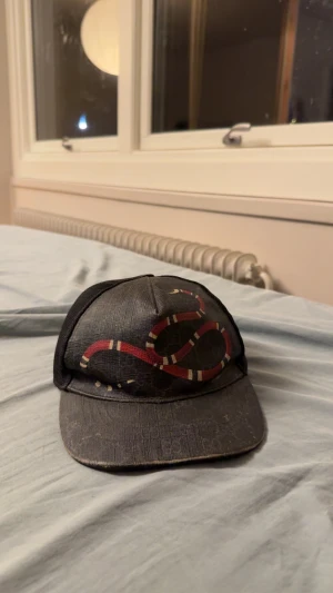 Svart Gucci keps med röd orm Äkta - Snygg svart keps från Gucci med mesh på sidorna och justerbar rem bak. Framtill finns ett coolt tryck med en röd, vit och svart orm som slingrar sig över kepsen. Materialet är polyester och kepsen har diskret GG-mönster i bakgrunden. De dåliga med kepsen är att den har slitits på lädret på visa ställen en till grej är att där man spänner kepsen är sönder. Kepsen är helt äkta👌🏽