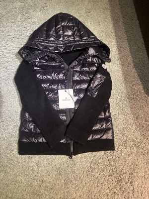 Svart Moncler pufferjacka med huva - Svart pufferjacka från Moncler med stickade ärmar och quiltad kropp. Jackan har en stor huva, dragkedja framtill och Moncler-logga på ärmen. Materialmix med polyester och stickad ull ger en schysst kontrast. Perfekt för dig som gillar streetwear och vill ha något som sticker ut, scan funkar. Hör gärna av er vid minsta fundering😊