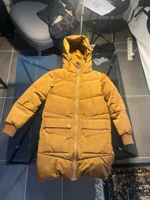 Senapsgul lång pufferjacka med huva - Lång pufferjacka i senapsgul färg med huva och ribbade muddar. Jackan har två stora fickor framtill med lock och knappar samt dragkedja hela vägen. Perfekt för kalla dagar och riktigt skön att bära. Reflex i fickan.