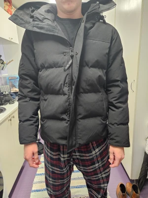 Svart pufferjacka - Säljer en svart pufferjacka med huva och justerbara snören. Jackan har lång ärm, dragkedja och ribbade muddar. Perfekt för kalla dagar med sin tjocka och quiltade design. Snygg och stilren look som funkar till de flesta outfits. Den är helt ny och oanvänd men har råkat ta bort prislappen. 