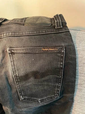 Svarta jeans från Nudie Jeans Co - Svarta jeans från Nudie Jeans Co i ekologisk bomull. Klassisk femficksmodell med svarta sömmar och diskret logga på bakfickan. Jeansen har en rak passform och snyggt slitna detaljer för en cool look. Hör gärna av er!