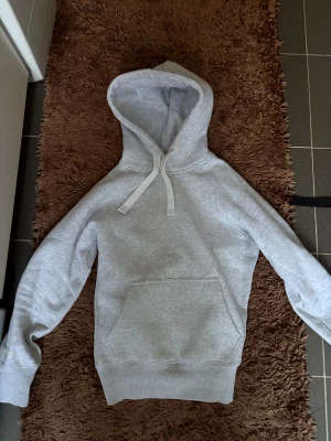 Russemerch - Snygg ljusgrå hoodie med klassisk huva och vita snören. Tröjan har en stor magficka framtill och ribbade muddar vid ärmslut och nederkant. Perfekt för chill dagar och enkel att matcha med jeans eller joggers.