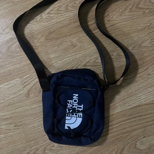 Mörkblå axelväska från The North Face - Snygg mörkblå axelväska från The North Face med vit logga på framsidan. Väskan har justerbar rem, dragkedja baktill och praktisk ficka framtill med elastiska snören. Perfekt storlek för mobil, plånbok och småprylar.