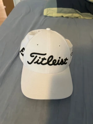 Vit Titleist keps Pro V1 - Snygg vit keps från Titleist med broderad svart logga framtill och Pro V1-text på sidan. Justerbar baktill med svart spänne och röd detalj runt loggan. Perfekt för golf eller chill på stan. Materialet är mjukt och lätt bomull. Tveka inte på att höra av er kring eventuella frågetecken kring pris, sick eller liknande!