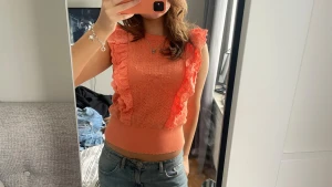 Korallfärgad blus med volanger - Superfin korallfärgad blus från Holly & Whyte med breda volanger längs axlarna och fram. Stickad struktur med hålmönster och broderade detaljer på volangerna. Croppad passform och rund hals. Perfekt för dig som gillar färg och coola detaljer.