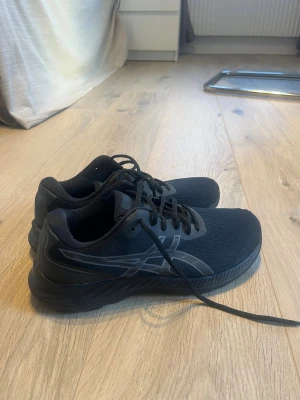 Svarta Asics sneakers strl 40.5 - Säljer ett par svarta Asics sneakers i storlek 40.5. Skorna har en sportig design med rund tå, platt sula och snörning. Ovandelen är i ventilerande syntetmaterial med diskret Asics-logga på sidan. Perfekta för dig som gillar stilrena och bekväma skor. Innermått 25,75 cm