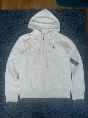Ljusgrå hoodie från Ralph lauren  - Vit hoodie från Polo Ralph Lauren med klassisk logga broderad på bröstet. Tröjan har huva med snörning, dragkedja framtill och två fickor. Tillverkad i mjuk bomullsmix, perfekt för chill dagar. Stilren och enkel design som funkar till allt.