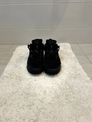 Svarta Nike Jordan 4 sneakers - Säljer ett par helsvarta Nike Jordan 4 sneakers med snygga detaljer och meshpaneler på sidorna. Skorna har snörning, platt sula och rund tå. Materialet är en mix av mocka och syntet, vilket ger en cool och sportig vibe. Perfekt för dig som gillar streetwear och vill sticka ut.