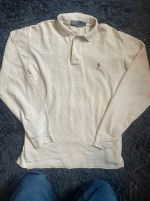 Beige långärmad pikétröja Polo Ralph Lauren - Snygg beige långärmad pikétröja från Polo Ralph Lauren i storlek M. Klassisk krage med två knappar och ikonisk broderad logga i vinrött på bröstet. Mjuk bomullskvalitet som är skön mot huden och passar perfekt till jeans eller chinos.