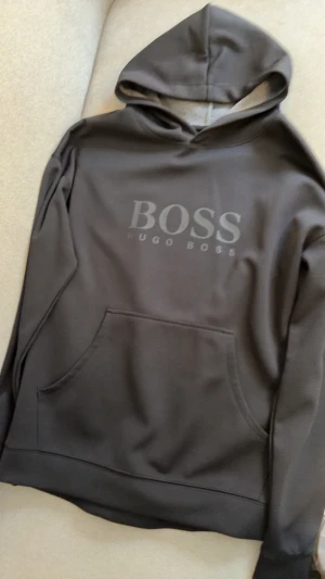 Mörkgrå hoodie från Hugo Boss - Snygg mörkgrå hoodie från Hugo Boss med stor BOSS-logga på bröstet. Tröjan har huva, lång ärm och en klassisk magficka framtill. Materialet är mjukt och känns som polyester, perfekt för chill dagar. Stilren och enkel design som funkar till många outfits.