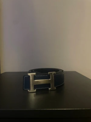 mörkblått skärp med H-spänne - Snyggt mörkblått bälte med ikoniskt H-spänne i metall. Bältet är gjort i skinn och har vita kontrastsömmar längs kanterna. Perfekt accessoar för att lyfta din outfit med en lyxig touch.