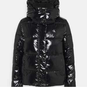Svart  vinterjacka  från Calvin Klein - Säljer en svart, glansig pufferjacka från Calvin Klein med  diskret CK-logga på bröstet. Jackan har långa ärmar, dragkedja och stora fickor framtill. Perfekt för dig som vill ha en snygg och varm jacka med streetkänsla.