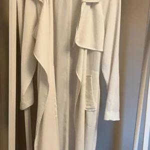 Vit omlott trenchcoat med fickor - Säljer en vit trenchcoat med snygg omlott-design och stora fickor framtill. Jackan har långa ärmar och ett avslappnat fall som ger en cool vibe. Perfekt för dig som gillar stilrena och enkla plagg med lite edge.