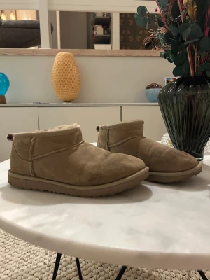 Beige UGG boots i mocka - Säljer ett par klassiska UGG boots i beige mocka med rund tå och platt sula. Insidan är fodrad med mjuk päls för extra värme och komfort. Skorna har en låg siluett och UGG-logga baktill. Perfekta för kalla dagar och chill stil.