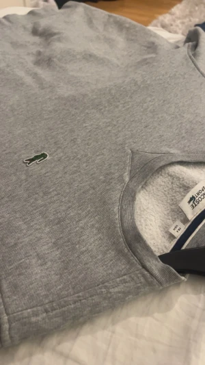 Grå sweatshirt från Lacoste Sport - Grå sweatshirt från Lacoste Sport med klassisk rund hals och det ikoniska gröna krokodilmärket broderat på bröstet. Mjuk insida och normal passform, perfekt för chill dagar. Enkel och stilren design som funkar till jeans eller joggers.