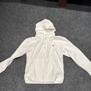 Vit hoodie från Polo Ralph Lauren - Säljs då jag gått upp några pannor och den är för liten                                                                         Snygg vit hoodie från Polo Ralph Lauren med klassisk broderad logga på bröstet. Tröjan har huva med snörning, dragkedja framtill och två stora fickor. Tillverkad i mjuk bomull, perfekt för chill dagar. Långärmad och stilren design.