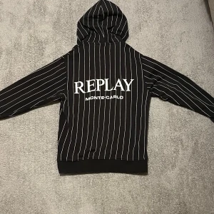 Svart randig hoodie Replay Monte Carlo - Svart hoodie från Replay med vita vertikala ränder och stor logotyp på ryggen. Framsidan har tryckt Replay-text och emblem, samt en klassisk känguruficka. Huva och ribbade muddar. Hoodien är väldigt liten i storlek och rekommenderas till folk med längden 152-165