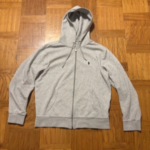 Grå hoodie från Polo Ralph Lauren - En snygg grå hoodie från Polo Ralph Lauren med dragkedja och huva med snörning som inte används längre utan bara sitter i garderoben. Den är bara använt fåtals gånger. Tröjan har två fickor fram och den klassiska broderade loggan på bröstet. Perfekt för chill dagar och enkel att matcha med jeans eller joggers. Vill få bort den så fort som möjligt!!!! Kan sänkas vid snabb affär!!!!