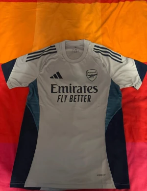Arsenal vit kortärmad fotbollströja - Snygg Arsenal fotbollströja från Adidas i vit med mörkblå detaljer och tryck. Tröjan har korta ärmar, rund hals och klassiska Adidas-ränder på axlarna. Materialet är lätt och andas, perfekt för träning eller match. Emirates Fly Better-logga på bröstet och Arsenal-emblem.